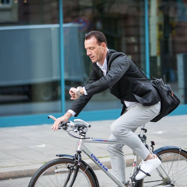 Mann fährt mit Fahrrad durch Stadt und guckt auf Uhr